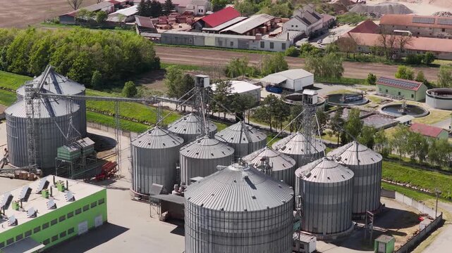 Aerial view of large metal grain silos at an industrial agricultural facility with surrounding green buildings and trees in Slavkov u Brna, South Moravian Region, Czechia.