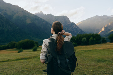 Fototapeta premium Young woman with a backpack gazes toward distant rugged mountains, ready for a hike, standing on a green meadow under a bright blue sky calm valley
