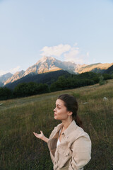 Fototapeta premium A young woman in casual beige jacket stands in a grassy field with mountains in the background under a clear sky. Lifestyle concept of nature exploration and outdoor relaxation.