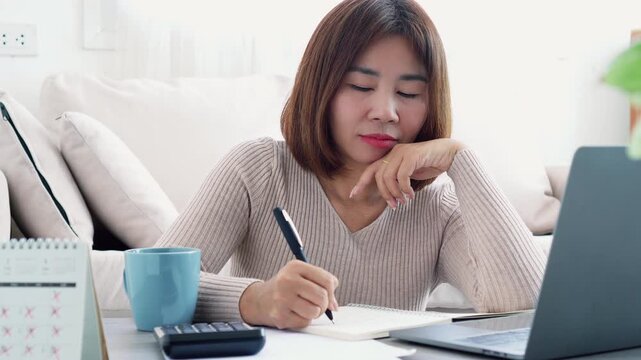Asian woman learning online, writing notes while focused on a laptop screen at home. Concept of e-learning, remote education, self-improvement, and professional development in a cozy workspace