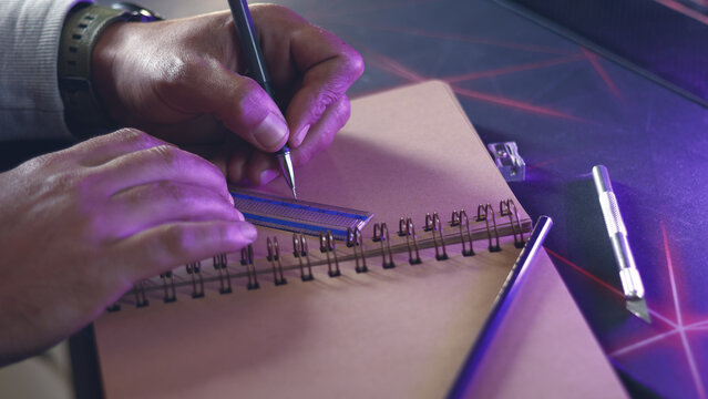 Left-handed designer drawing with pen and ruler in sketchbook under purple and blue neon lighting