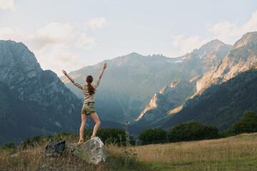 Fototapeta premium Young woman outdoors lifestyle on a rock with arms raised in a scenic mountain landscape, celebrating nature, hiking, freedom, and adventure under bright daylight.