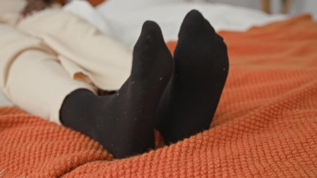 Woman in black socks lying on bed with ankles crossed on an orange blanket in studio; serenity cozy rest.