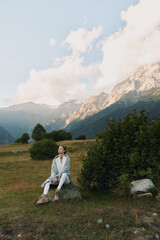 Fototapeta premium Young woman in a light denim jacket and jeans sits on a rock in a green meadow, with distant alpine mountains and a bright sky forming a dramatic natural backdrop
