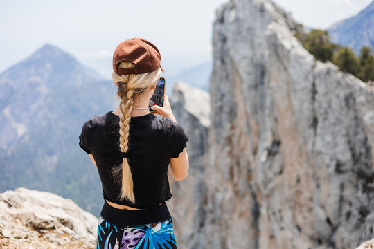 Woman stands on rocky mountain peak, taking photo with smartphone