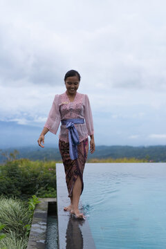 Smiling Balinese woman in kebaya walking along infinity pool, Bali