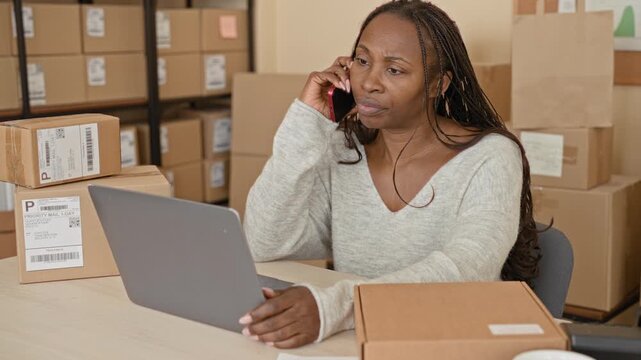 Woman holding smartphone to ear while typing on laptop amid stacked shipping packages in a building; concern entrepreneurship.