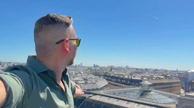 Smiling man filming himself on a sunny day from a viewpoint overlooking the beautiful city of Paris, with the famous Palais Garnier Opera House visible in the background