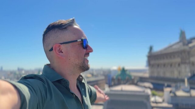 Young man wearing sunglasses smiling and filming himself on a rooftop while enjoying the beautiful cityscape of Paris, France on a sunny day with clear blue sky during his european vacation