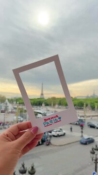 Personal perspective of a female hand holding a Bonjour Paris souvenir frame, capturing the iconic Eiffel Tower and the bustling city skyline from a high angle view during a vacation