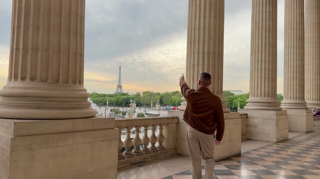 Young male tourist standing on a balcony with large columns admiring the spectacular view of the Eiffel Tower and the Paris skyline, pointing at the famous landmark during his vacation