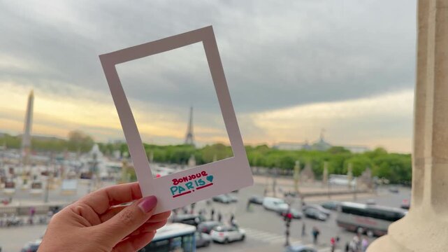 Close-up of a female hand holding a paper frame with the text Bonjour Paris over the famous Place de la Concorde, framing the Eiffel Tower and the Obelisk of Luxor during a beautiful sunset