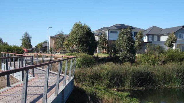 Boardwalk over wetland reserve beside modern homes in Point Cook, Melbourne, Australia. People biking through neighborhood parkland. Green infrastructure, active lifestyle amenity