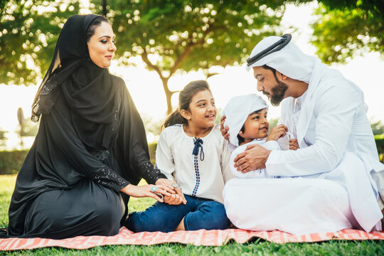 Arabian family in traditional dress smiling together on a picnic blanket in a sunny green park, parents and children enjoying leisure, bonding and relaxation outdoors