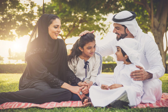 Arabian family in traditional dress smiling together on a picnic blanket in a sunny green park, parents and children enjoying leisure, bonding and relaxation outdoors
