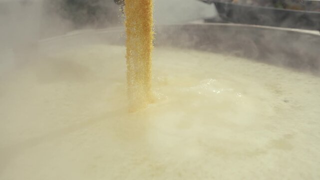 Person pouring fine yellow powder into a large pot of boiling liquid, steam rising, showcasing the continuous action of mixing ingredients in a cooking process