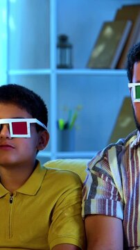 Indian young family watching 3d movie at home wearing goggles and enjoying popcorn together while sitting on sofa indoors during fun movie night inside a modern lavish living room with joy