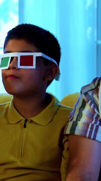 Indian young family watching 3d movie at home wearing goggles and enjoying popcorn together while sitting on sofa indoors during fun movie night inside a modern lavish living room with joy