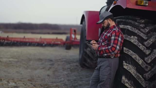 Farming and agribusiness, farmworker standing near tractor and using smartphone. Professional tractor driver resting after long hard working day in farmland, online shopping in mobile application