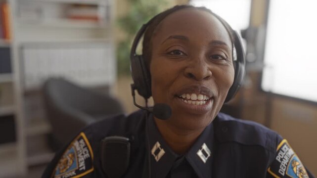 Young african american woman police officer wearing headset and radio speaking and smiling with mouth visible inside building; duty trust reassuring.