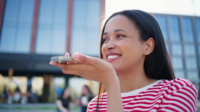 Close up view of woman sending audio message on smartphone outdoors. Female speaking near mobile device while relaxing in downtown area. Person using digital communication during daytime.