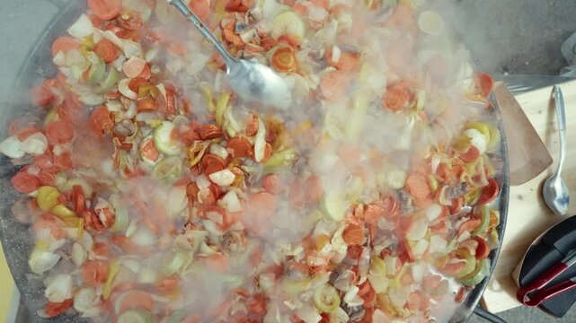 Chef in black gloves stirs colorful mixed vegetables in a large pan, steam rising, showcasing vibrant ingredients in an outdoor cooking setting