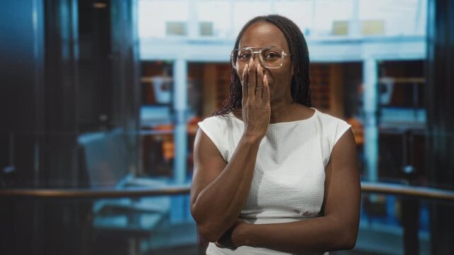 Young african american woman in white dress hand to mouth smiling wearing clear glasses and arm crossed near railing in building atrium; surprise warmth.