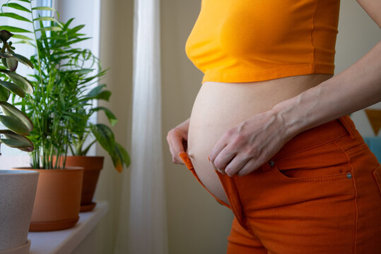Pregnant woman struggles to fasten her outgrown orange jeans, showing her expanding belly and the common experience of body changes and weight gain during pregnancy, needing new maternity wear