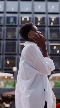 Black woman talking on mobile phone while walking on city street.backdrop of contemporary architecture and busy city street.Female enjoy mobile phone conversation, confident and professional demeanor

