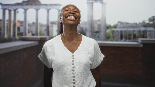 Woman in white button blouse smiling and giving thumbs up showing thumb and teeth on a rooftop building terrace with brick parapet and ancient columns; joy confidence.