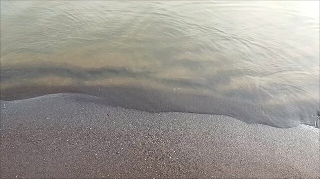 Gentle waves washing over dark sandy beach shoreline with calm water ripples, creating a peaceful tropical coastal atmosphere. Perfect natural background for relaxation and seaside themes