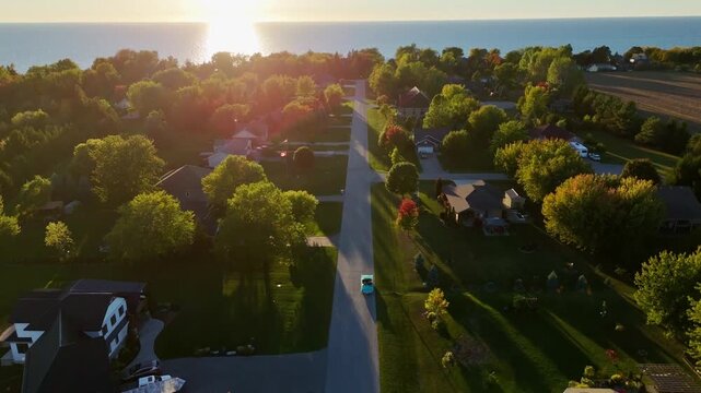 Cinematic drone shot following a vintage car driving through a quiet suburban neighborhood towards a golden Lake Huron sunset. Beautiful evening glow in Ontario.