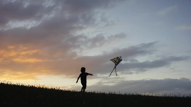 Child running joyfully on grassy hill at sunset, flying colorful kite with vibrant tail, silhouetted against dramatic sky filled with clouds and warm hues