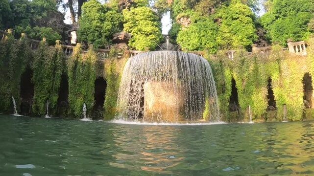 Tilt down reveals grand oval fountain cascading into pool, surrounded by lush greenery and ancient stone niches.