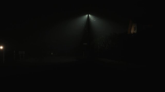 Camera moving forward along foggy night road toward streetlight, smooth motion enhances atmosphere of empty street with dense fog dim lighting and strong sense of low visibility