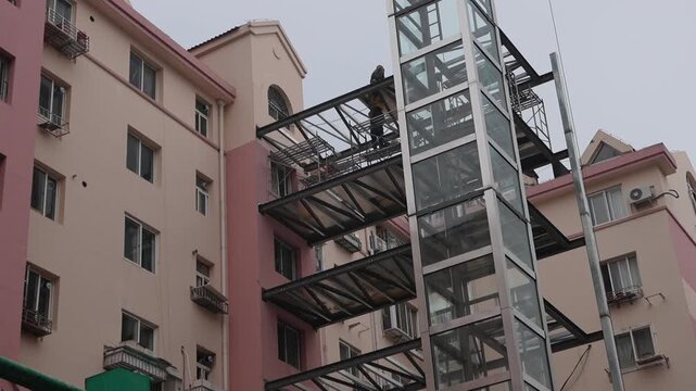 This photo shows outdoor elevator renovation construction on an old residential apartment building, with workers assembling the steel and glass elevator frame and attached balconies.