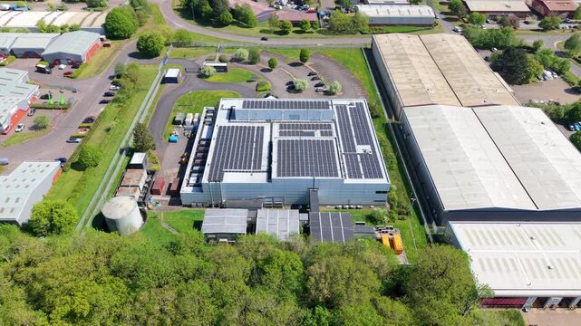 Aerial view of a modern data center building featuring solar panels on the roof surrounded by lush green trees and industrial warehouses in Wellingborough, England, United Kingdom.