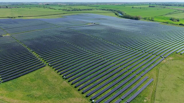 Aerial view of a large solar farm with rows of photovoltaic panels surrounded by green fields and trees in Irchester, England, United Kingdom.
