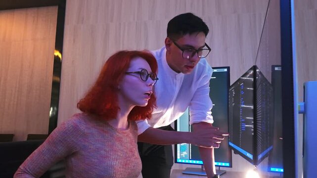 Two colleagues in glasses lean toward glowing monitors, reviewing code late in a modern office, reacting with focus and surprise.