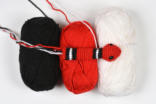 Top view of a small handmade crocheted milk snake toy lying horizontally across black red and white yarn skeins on a plain background.