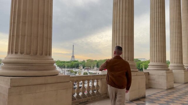 Young man wearing a jacket, walking on a balcony with large columns and pointing at the Eiffel Tower in the distance, enjoying the magnificent cityscape view of Paris from the Trocad&eacute;ro