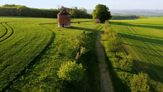 Aerial view of the historic brick windmill standing in lush green fields with rolling hills and a tree-lined path in Chvalkovice, Hradec Kralove Region, Czechia.