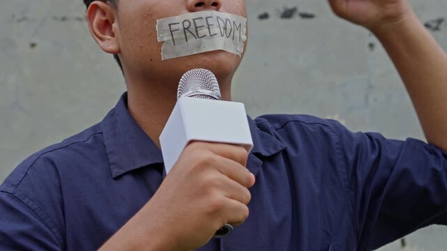 Close-up of silenced reporter with "FREEDOM" tape on his mouth holding microphone and raising fist