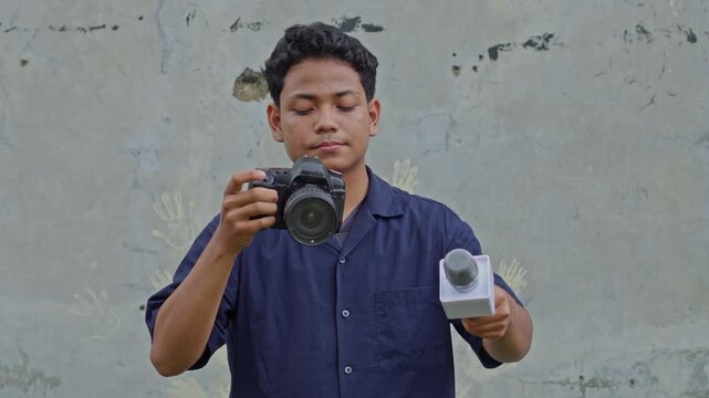 Young male journalist holding DSLR camera and professional microphone against concrete wall