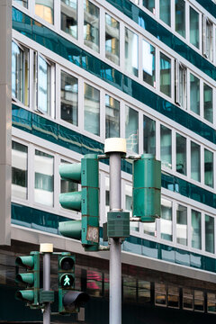 an urban green traffic light fixture and crosswalk signal pole rise along a street, set against a building facade of windows and columns