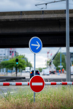 a traffic barrier and blue arrow sign align near grass, holding road direction and signal layers across a quiet urban street edge.
