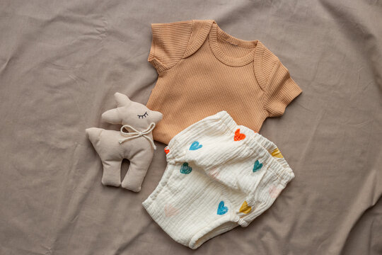 Beige baby bodysuit and white cotton diaper pantie with toy on beige sheet, top view. Eco friendly cloth and accessories