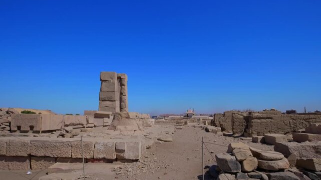 Two ruined ancient Egyptian granite columns standing on Elephantine Island ruins, Aswan, Egypt, with scattered stone blocks, fallen capital and city skyline under vivid blue sky.