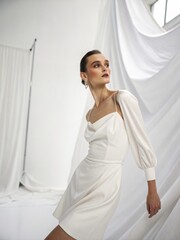 Fototapeta premium High Fashion Romantic Portrait of Elegant Woman in Silk Ivory Dress against Flowing Fabric Drapes and Soft Light Perfect for Bridal, Luxury Product or Cloud Dancer Color Advertising