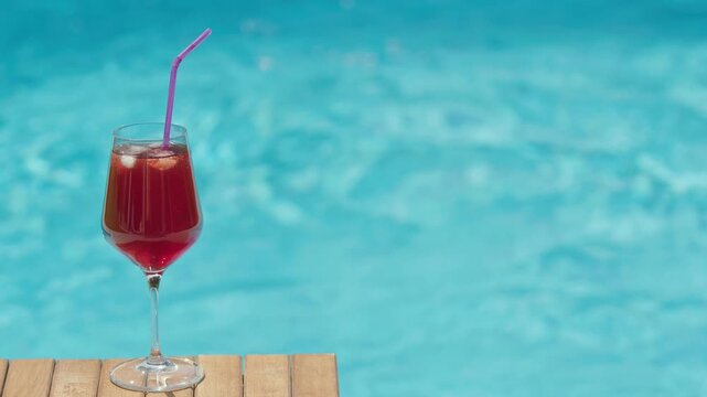 Red cocktail with ice and straw standing on wooden table by swimming pool water on sunny day.